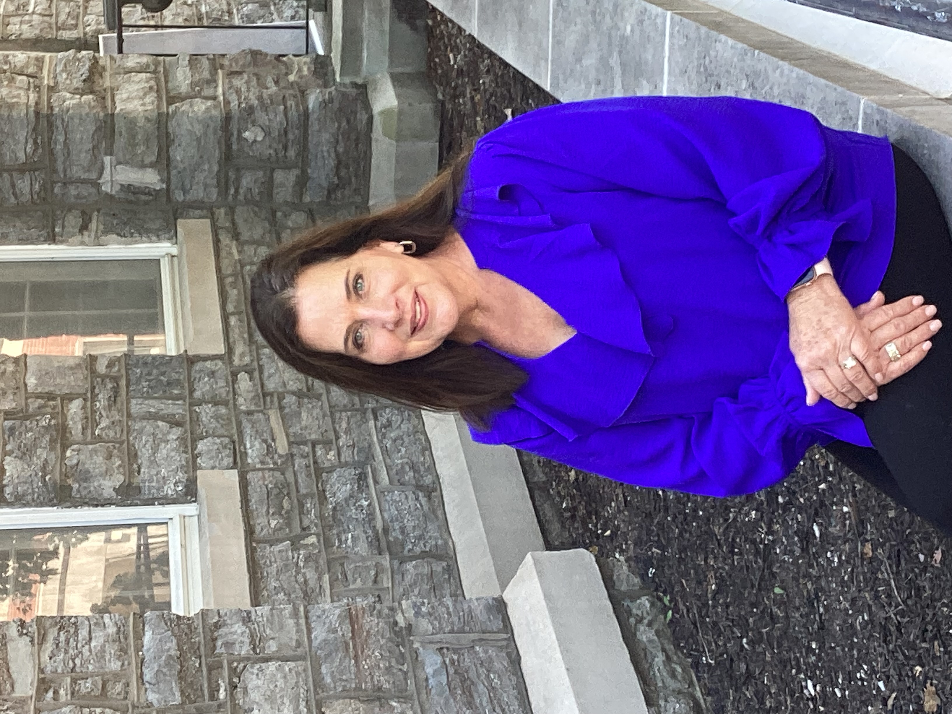 Woman with long brown hair in a bright purple blouse sitting on a stone bench in front of a brick building